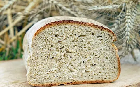 Ein frisch gebackenes, aufgeschnittenes Brot liegt auf einem Holzbrett. Im Hintergrund sind Ähren zu sehen, die an ein Kaninchen erinnern, das sich am Brot labt.