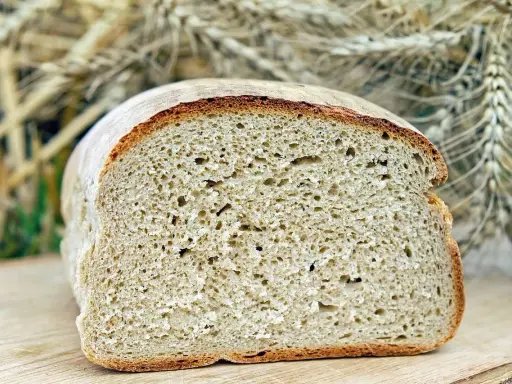 Ein frisch gebackenes, aufgeschnittenes Brot liegt auf einem Holzbrett. Im Hintergrund sind Ähren zu sehen, die an ein Kaninchen erinnern, das sich am Brot labt.