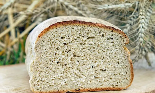 Ein frisch gebackenes, aufgeschnittenes Brot liegt auf einem Holzbrett. Im Hintergrund sind Ähren zu sehen, die an ein Kaninchen erinnern, das sich am Brot labt.